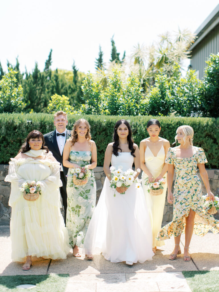A bride and five bridesmaids in floral dresses and holding bouquets walk outside on a sunny day.