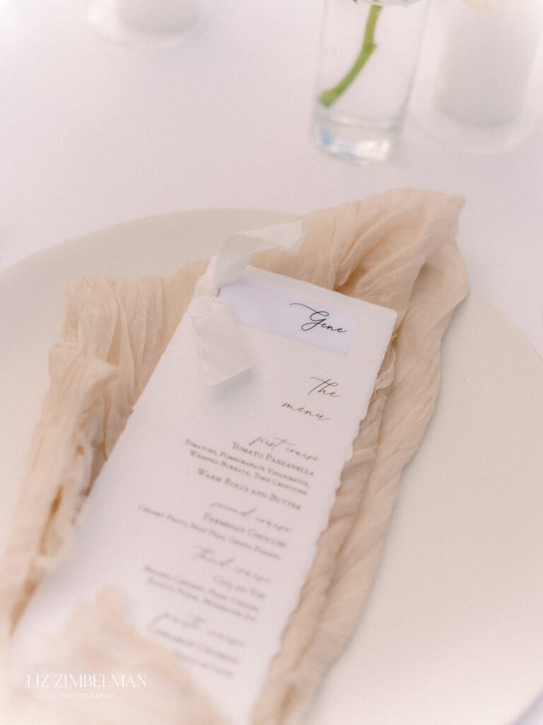A restaurant table setting with a menu placed on a cream-colored cloth napkin on a white plate. A small white bow holds the menu. A flower in a vase is partially visible in the background.