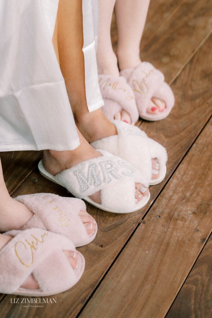 Close-up of three people wearing fluffy slippers. One pair is white with "Mrs" in rhinestones, and the other two are pink with gold lettering. All stand on wooden flooring.