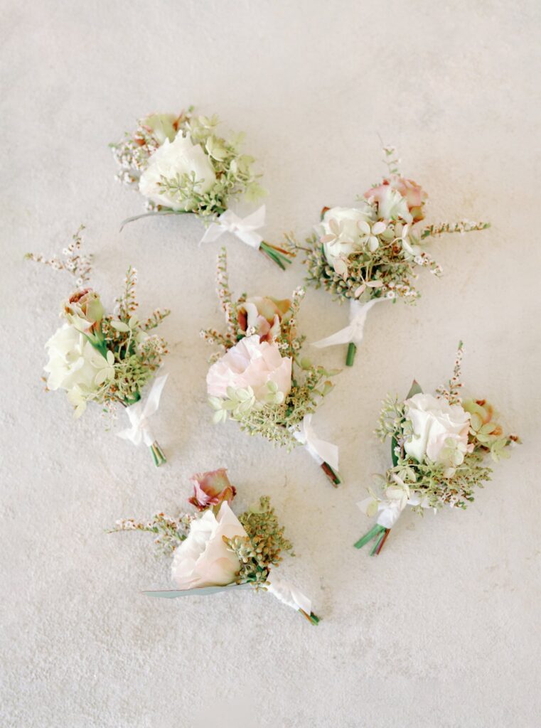 Six small floral boutonnieres with white and pink flowers and greenery arranged on a light textured background.