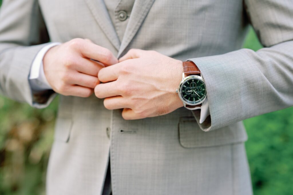 A person in a light gray suit adjusts their jacket. They wear a watch with a leather strap on their left wrist.