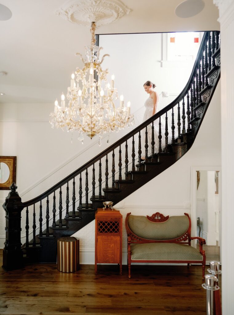 A person in a white outfit descends a dark wooden staircase in an elegantly decorated room with a chandelier and vintage furniture.