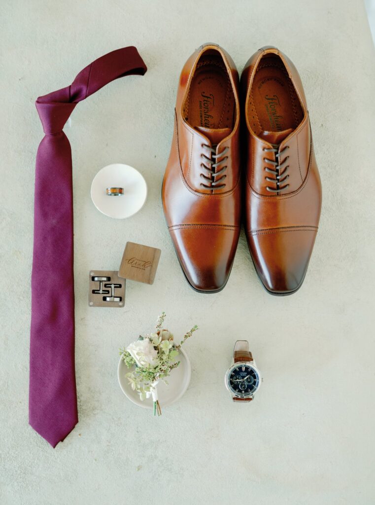 Brown leather dress shoes, a burgundy tie, cufflinks, two rings in small dishes, a wristwatch, a wooden box, and a small floral arrangement on a light surface.