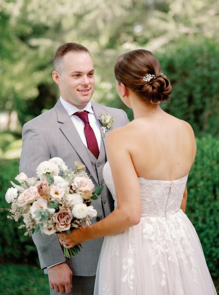 A bride and groom stand facing each other outdoors. The bride holds a bouquet of flowers and wears a strapless white dress. The groom wears a gray suit with a burgundy tie.