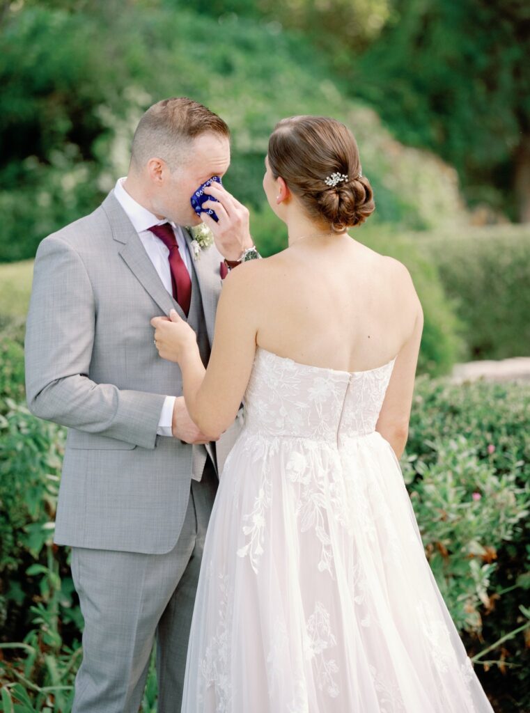 A bride in a white dress stands with a groom in a gray suit who is wiping his face with a blue cloth, surrounded by greenery.
