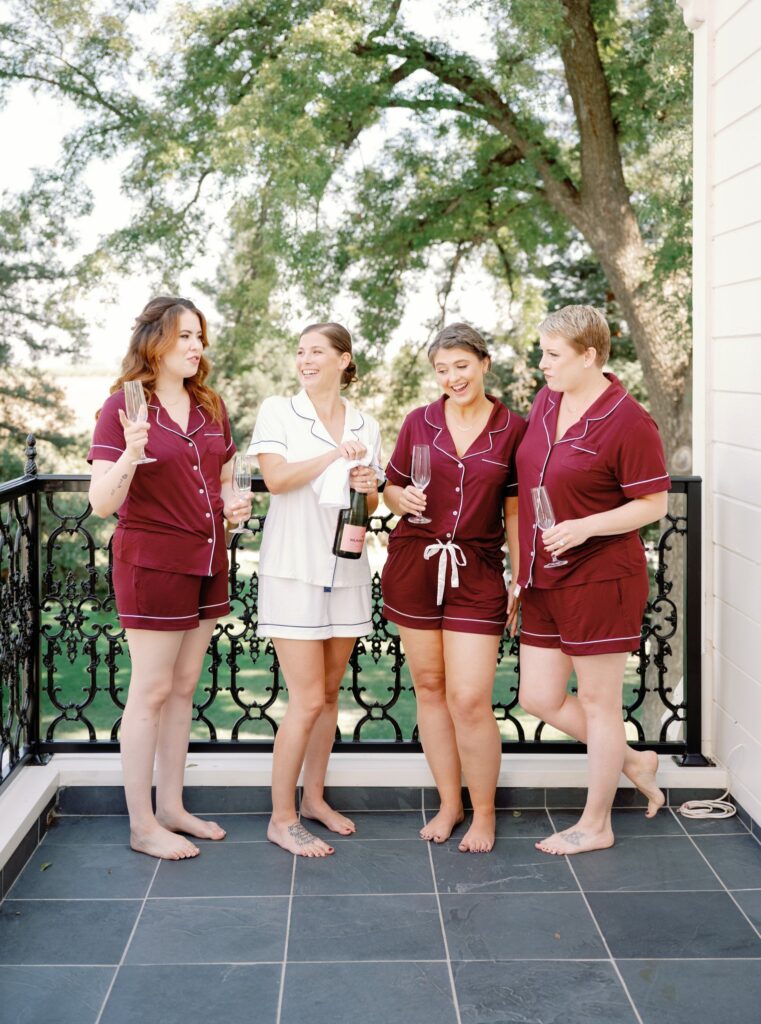 Four women in pajamas stand barefoot on a balcony, holding champagne flutes and a bottle, surrounded by trees in the background.