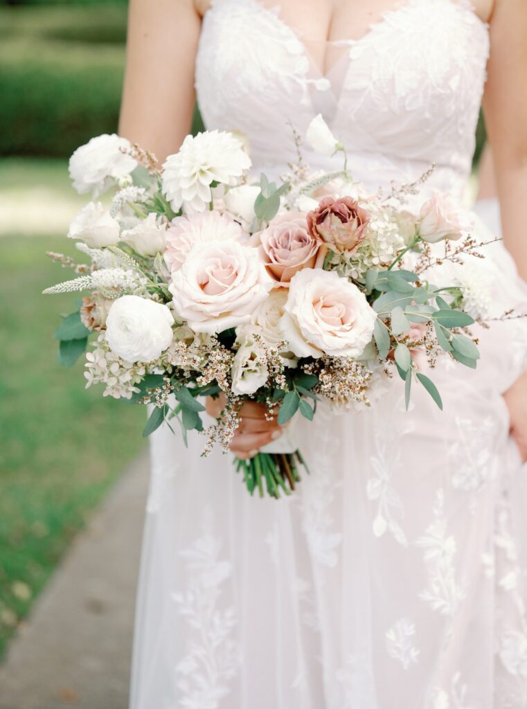 Bride holding a bouquet of white and pink flowers with greenery, wearing a white lace dress in an outdoor setting.