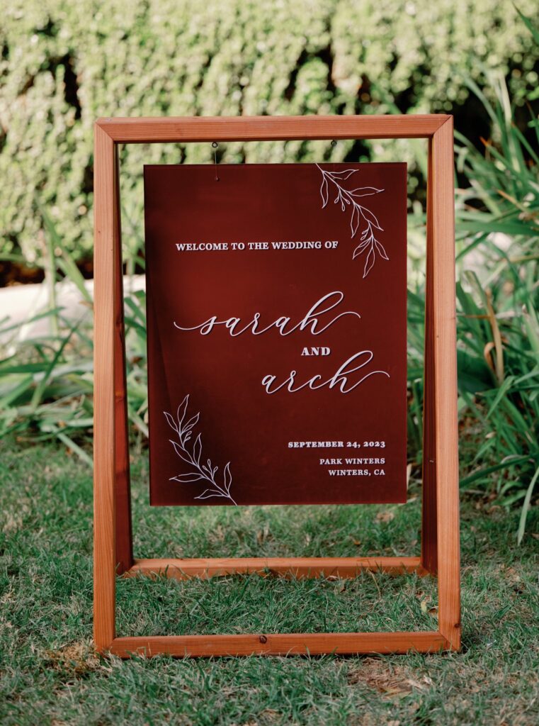 Wedding welcome sign for Sarah and Arch, featuring floral designs, held in a wooden frame on grass.