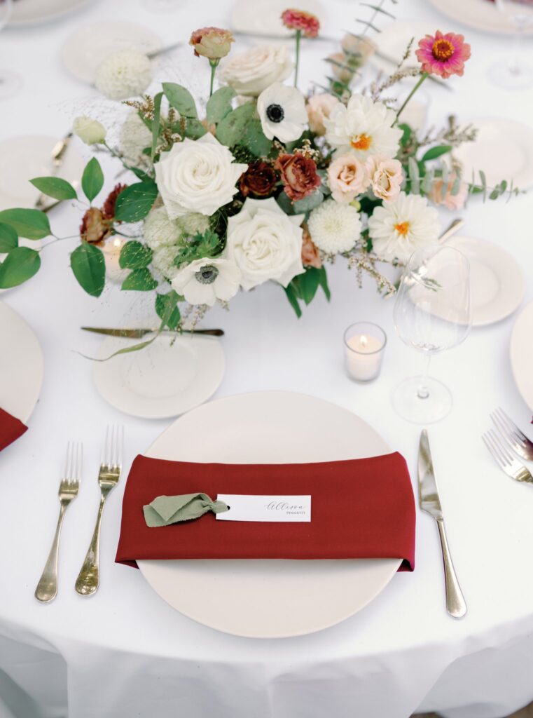 A round table set with a floral centerpiece, white plates, gold cutlery, a wine glass, and a red napkin with a name card.