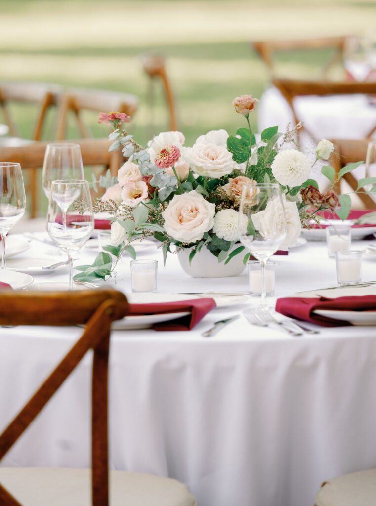 A round table is set for an event with a white tablecloth, floral centerpiece, wine glasses, and folded red napkins on plates. Wooden chairs surround the table.