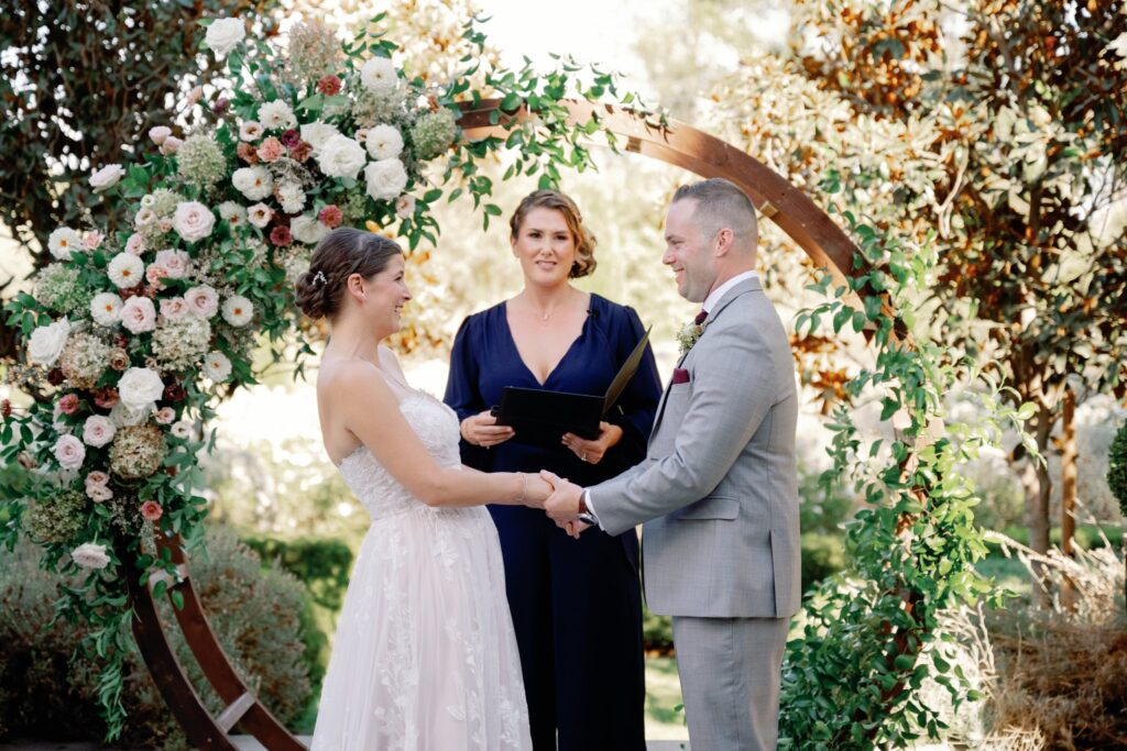 A couple holds hands during their wedding ceremony in front of a floral arch, with an officiant standing between them.