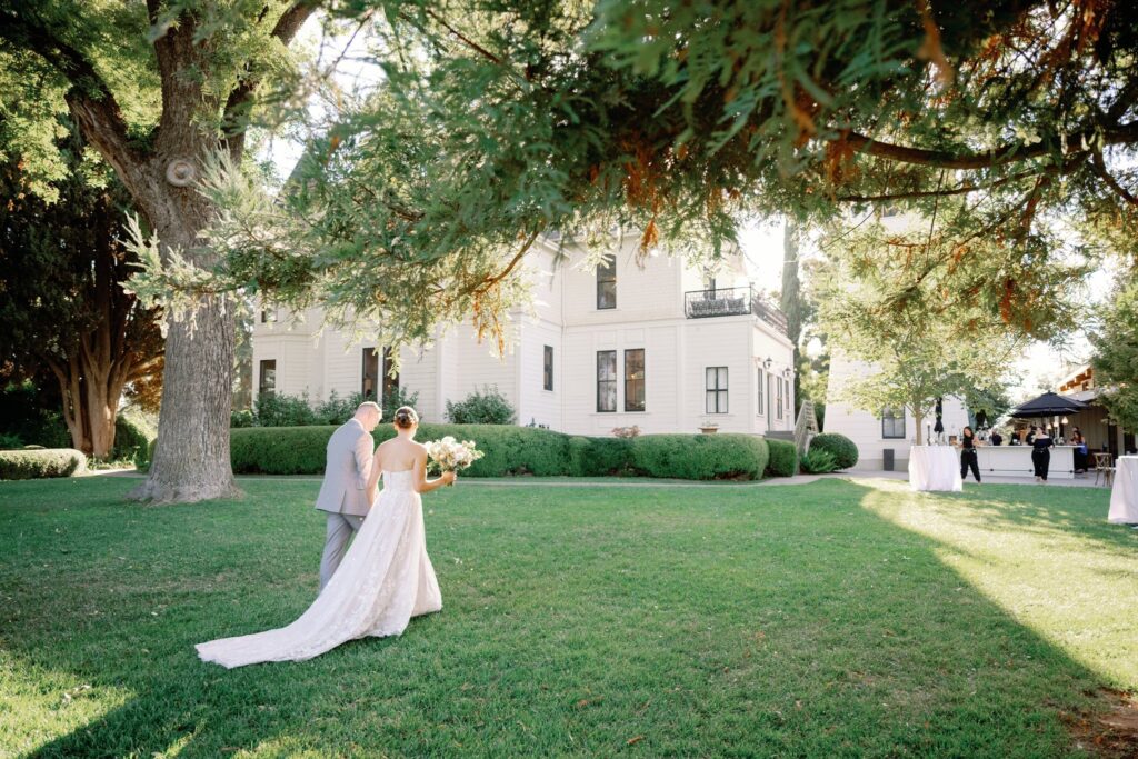 A bride and groom walk across a grassy lawn toward a large white house, surrounded by trees, on a sunny day.
