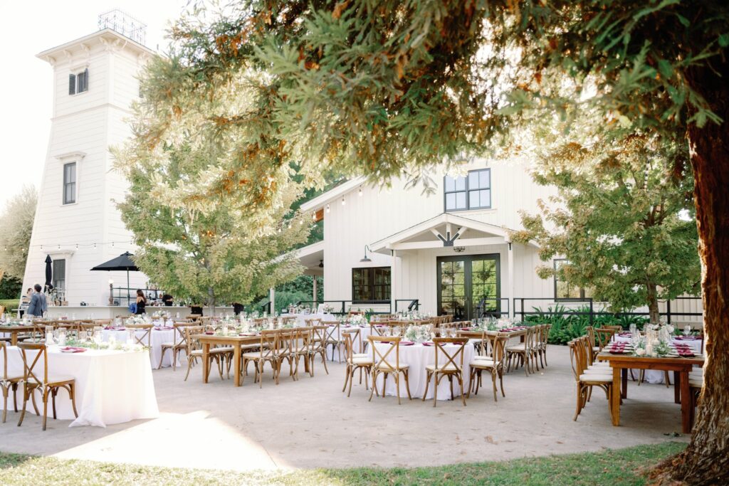 Outdoor wedding reception setup with wooden tables and chairs on a patio, surrounded by trees, near a white barn-like building and a tower.