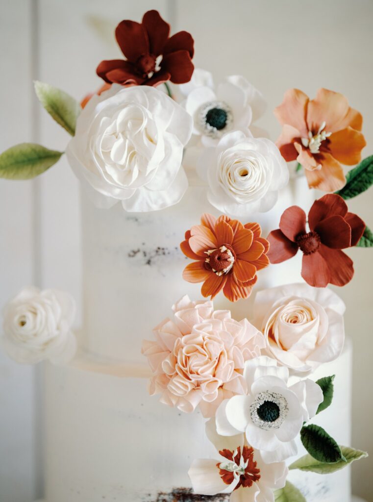 A white tiered cake adorned with realistic-looking edible flowers in shades of white, pink, and red, with green leaves.