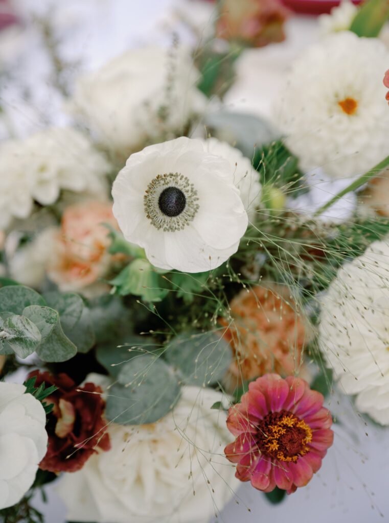 Close-up of a floral arrangement featuring white anemones, pink and red zinnias, and green foliage on a light background.