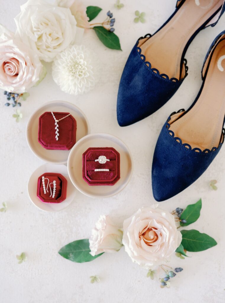 Flat lay of rose flowers, navy blue scalloped shoes, and three open red velvet jewelry boxes displaying a necklace, earrings, and a ring set on a light surface.