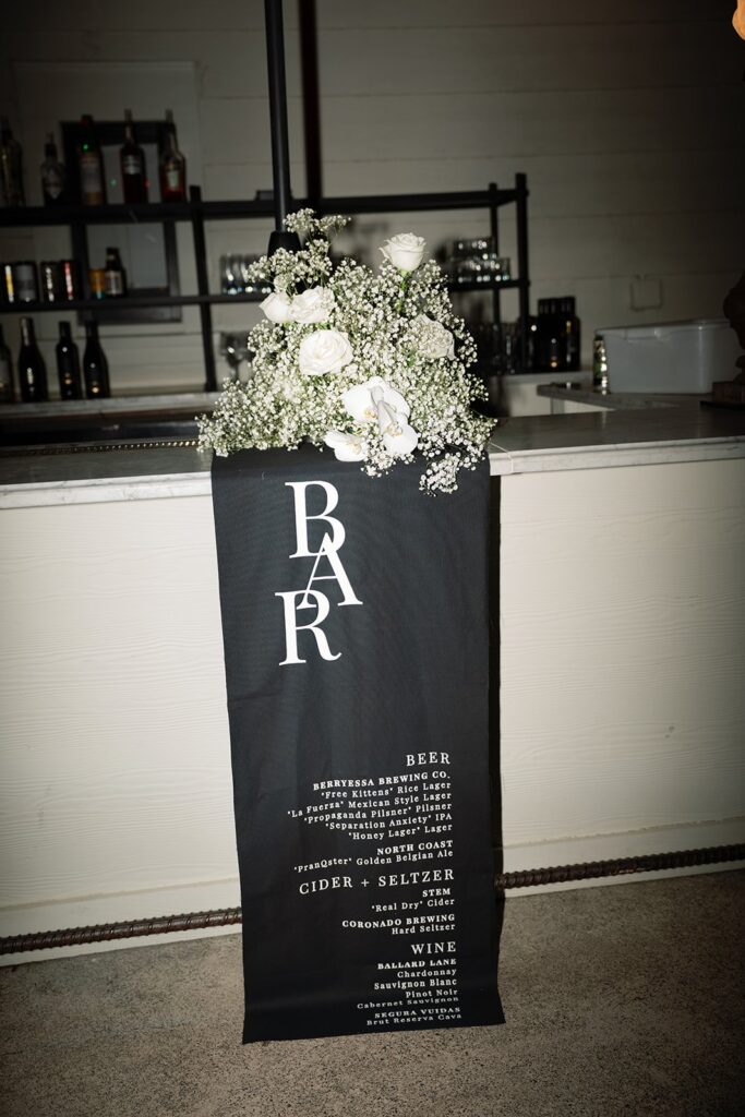 A black bar menu sign with white text stands in front of a bar counter, adorned with white flowers and greenery at the top.
