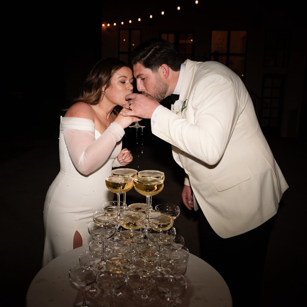 A couple in formal attire drinks from glasses at the top of a champagne tower.
