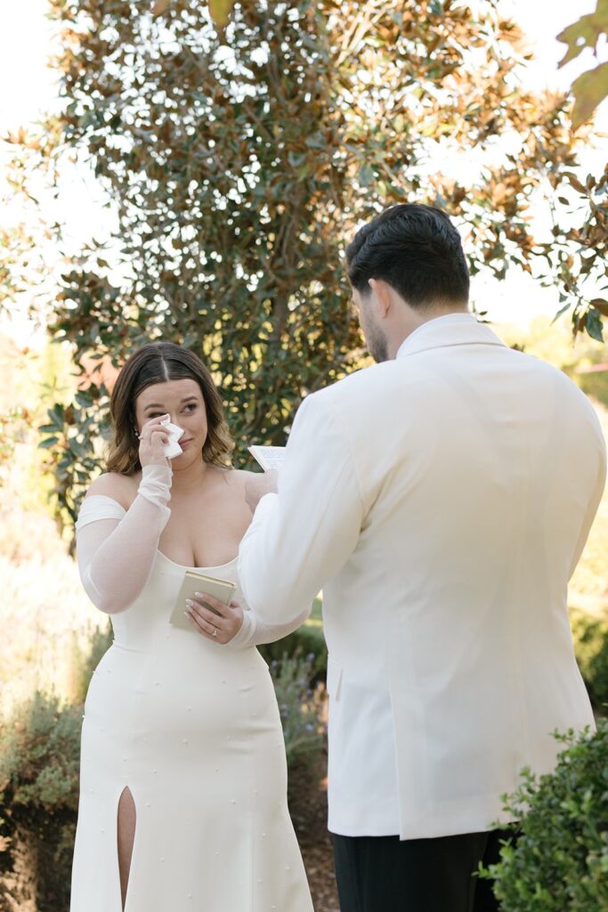 A bride wipes her tears while the groom reads vows during an outdoor ceremony. Both are dressed in white.