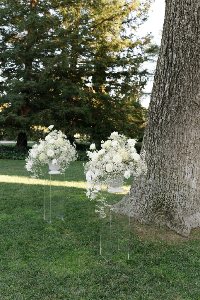 White floral arrangements on stands are placed on grassy ground next to a large tree trunk, with trees in the background.