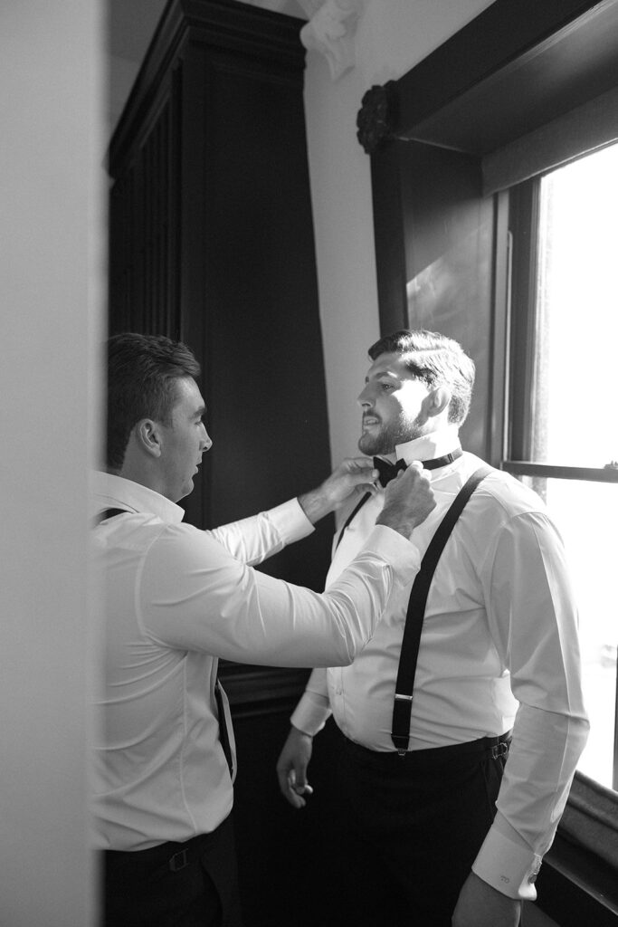 Two men in dress shirts and suspenders, one adjusting the other's bow tie, standing near a window with light streaming in.