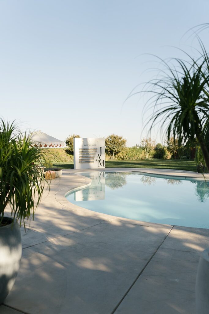 Outdoor swimming pool with surrounding greenery and a white building in the background. Two large potted plants are in the foreground.