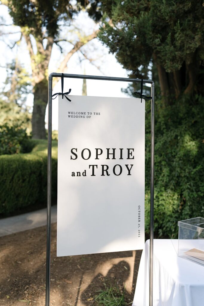 A sign at an outdoor wedding reads "Welcome to the wedding of Sophie and Troy." It is displayed on a metal stand with trees in the background.