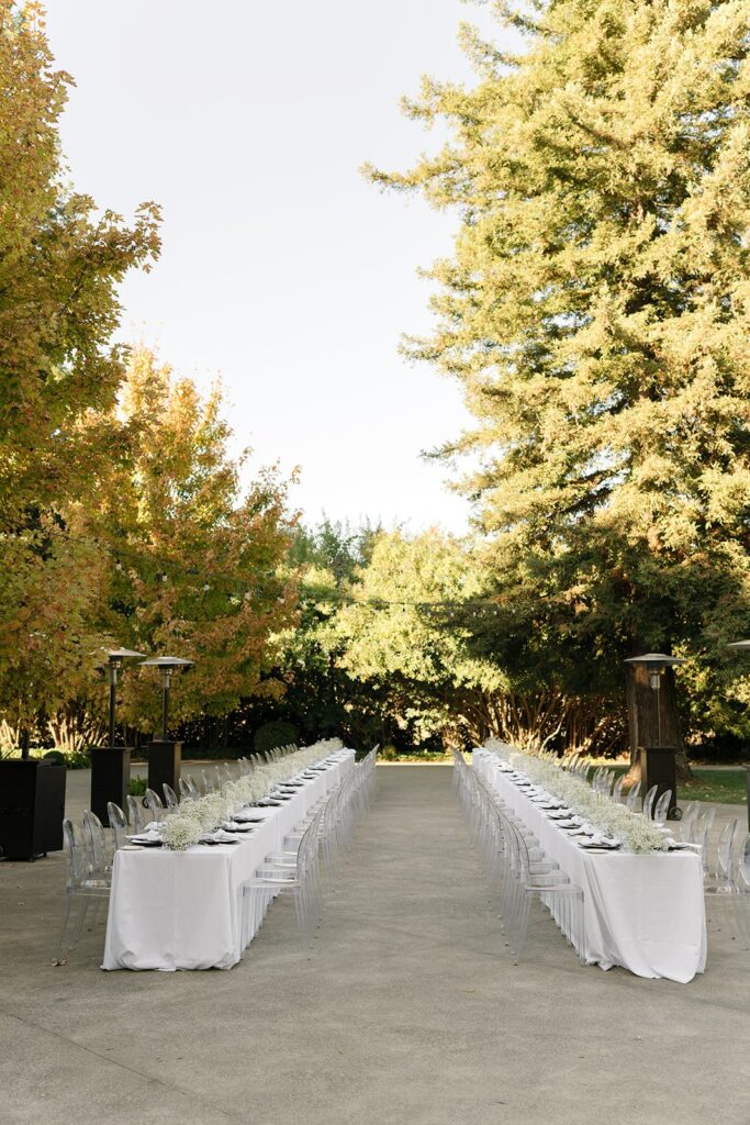Long outdoor dining setup with two rows of tables covered in white tablecloths and arranged with place settings, surrounded by lush green trees.