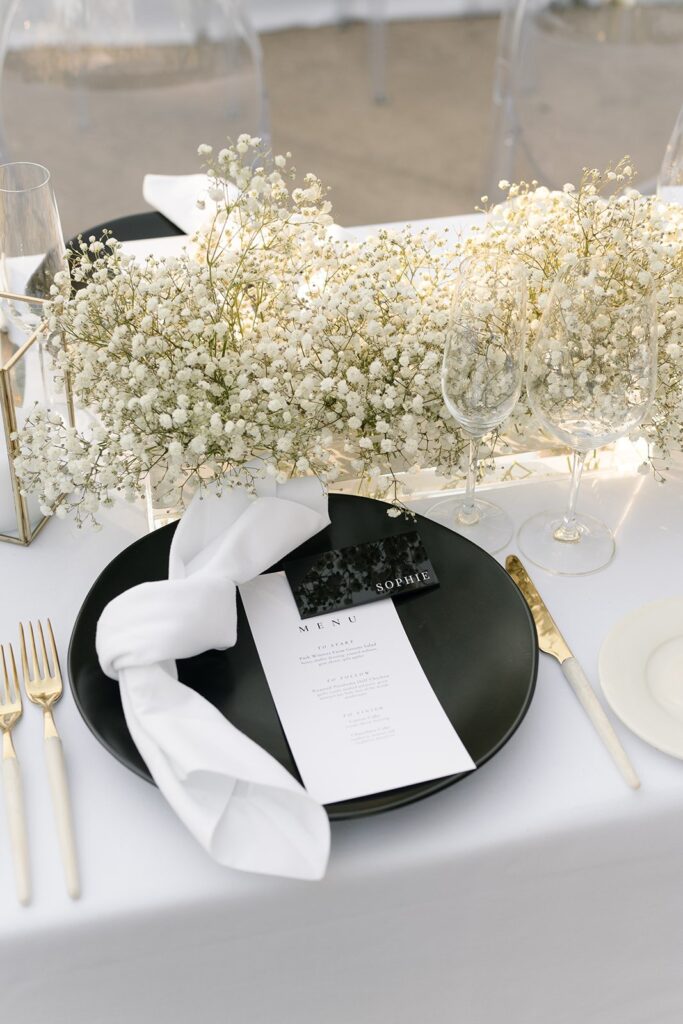 Elegant table setting with a black plate, white napkin, menu, and gold utensils. Delicate white flowers and empty wine glasses are arranged on a white tablecloth.