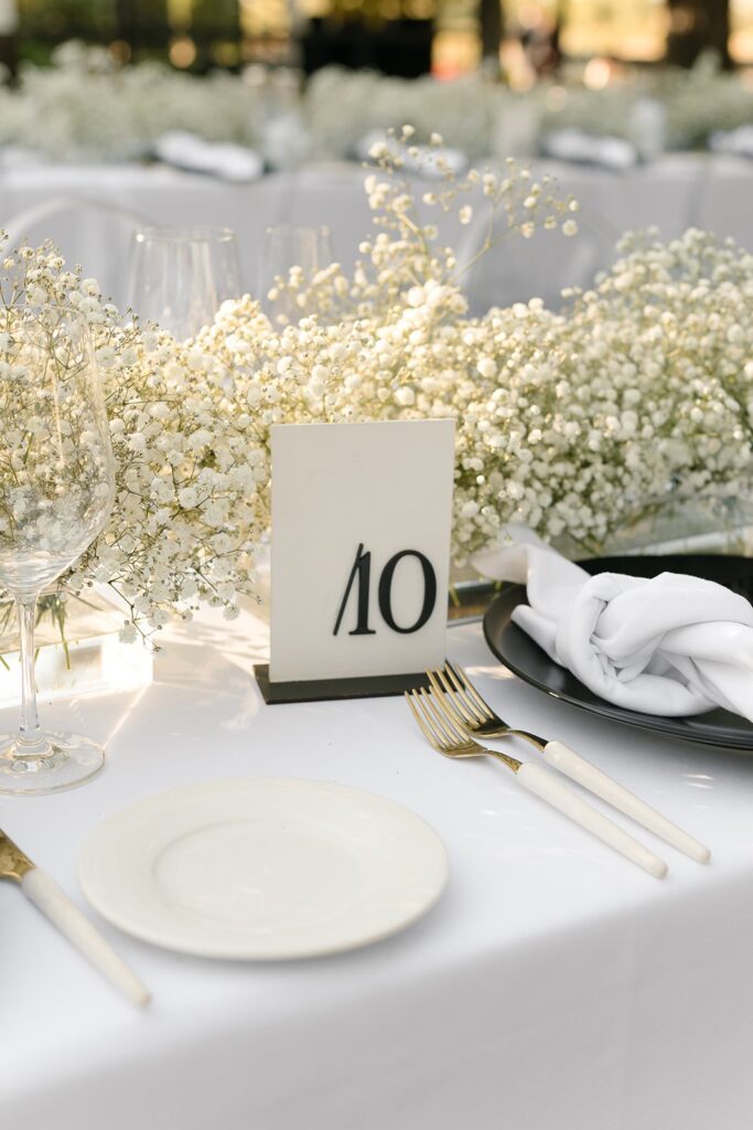 Table setting with a white tablecloth, black plate, white napkin, and gold cutlery. A floral centerpiece with baby's breath surrounds a table number card displaying "10.
