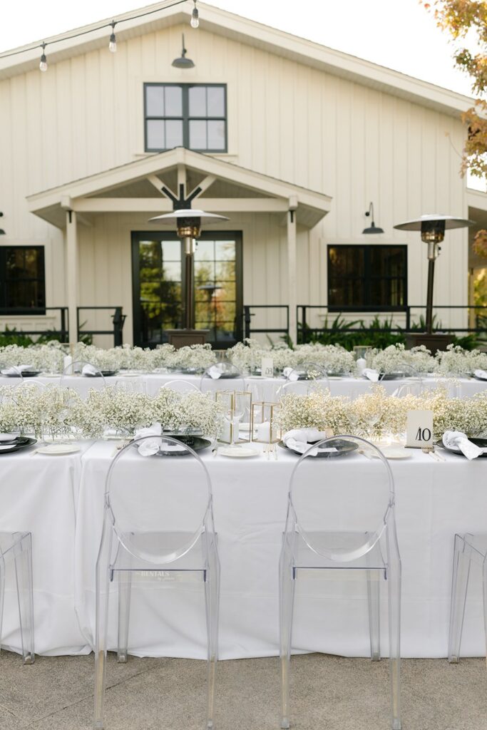Outdoor event setup with white tablecloths and clear chairs in front of a white building. Tables are decorated with white flowers and candles.