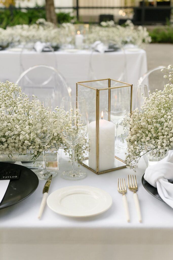 Elegant table setting with white dishes, cutlery, and wine glasses. A lit candle in a glass holder and white flowers decorate the center. Transparent chairs and a white tablecloth are visible.