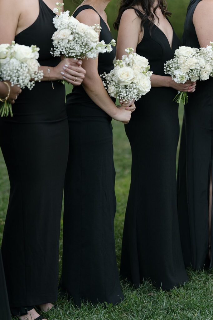 A row of people in black dresses holding white rose and baby's breath bouquets, standing on grass.
