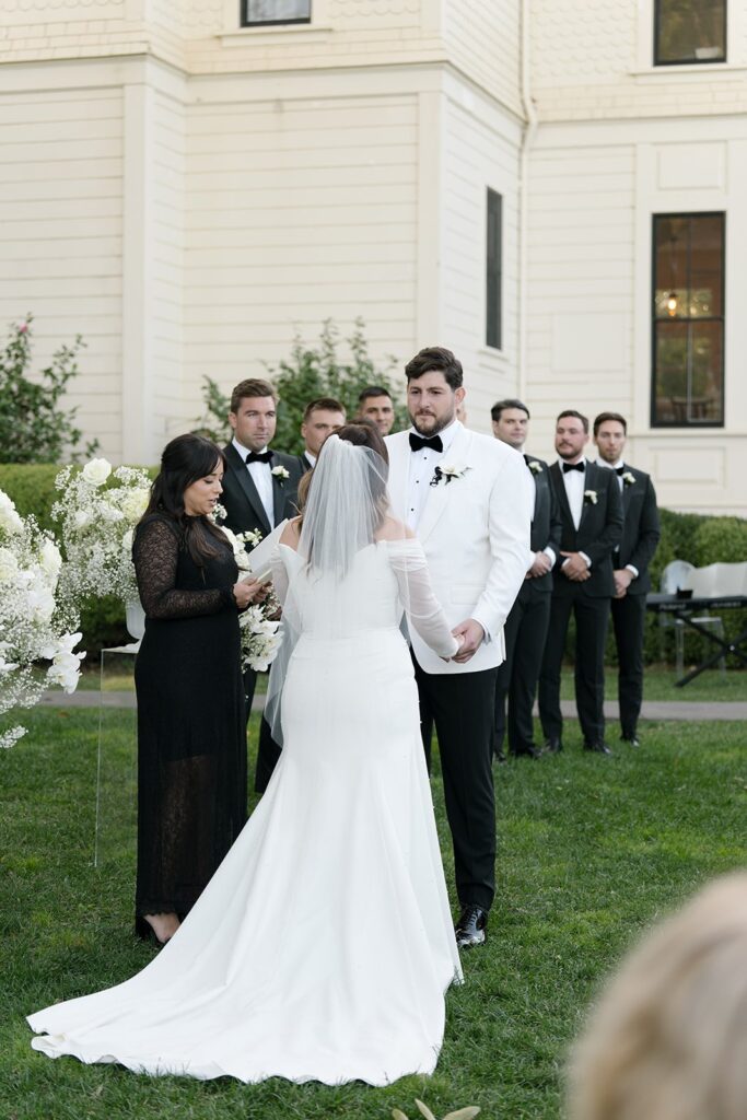 A couple stands in a garden during their wedding ceremony, exchanging vows. The bride wears a white gown and veil, while the groom wears a white tuxedo jacket. Bridesmaids and groomsmen stand nearby.