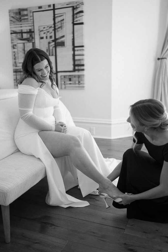 A woman in a gown sits on a sofa, smiling, as another woman helps her with her shoe in a room with modern decor.