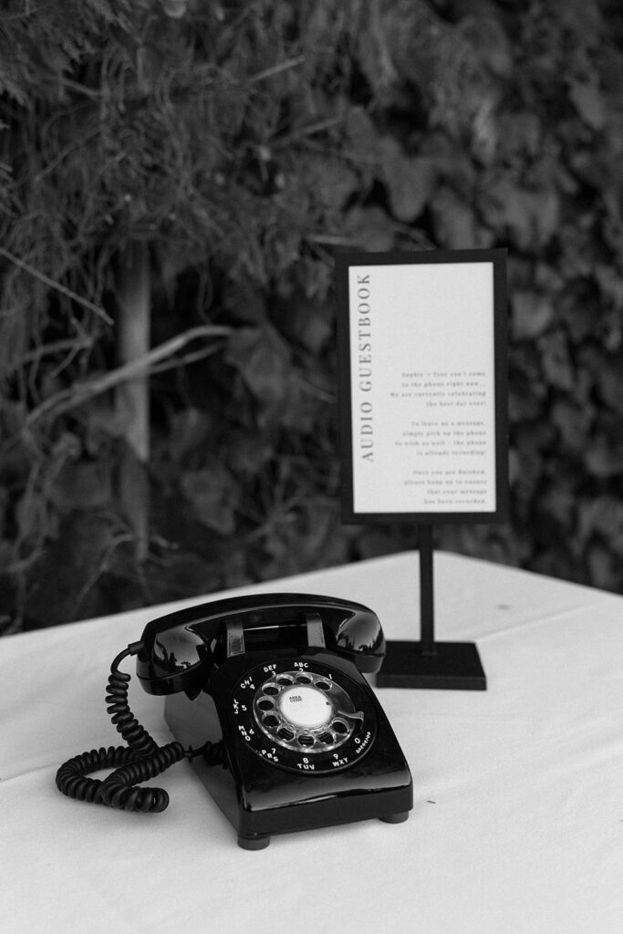 A vintage rotary phone sits on a table next to a sign labeled "Audio Guestbook," set against a backdrop of foliage.