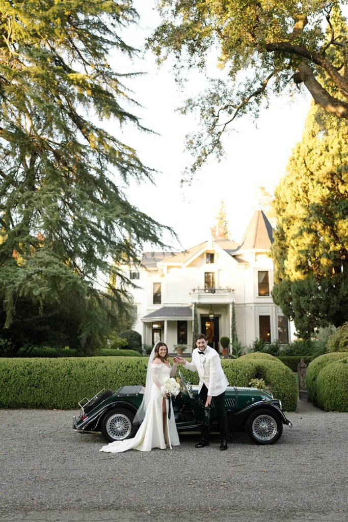 A couple in wedding attire stands beside a vintage black car in front of a large white house surrounded by greenery.