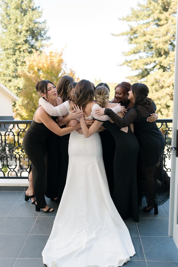 A group of women in black dresses hug a woman in a white dress on a balcony with trees in the background.