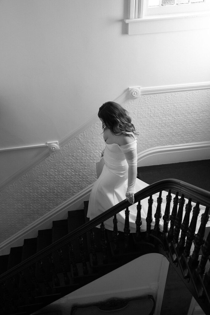 A woman in a white off-shoulder gown descends a staircase, her hand resting on the railing. The setting is indoors with minimal light.