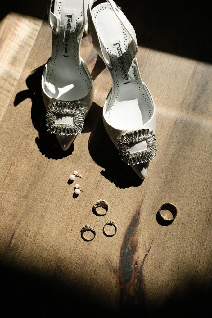 High-heeled shoes with jeweled buckles are placed on a wooden surface alongside pearl earrings and three rings.