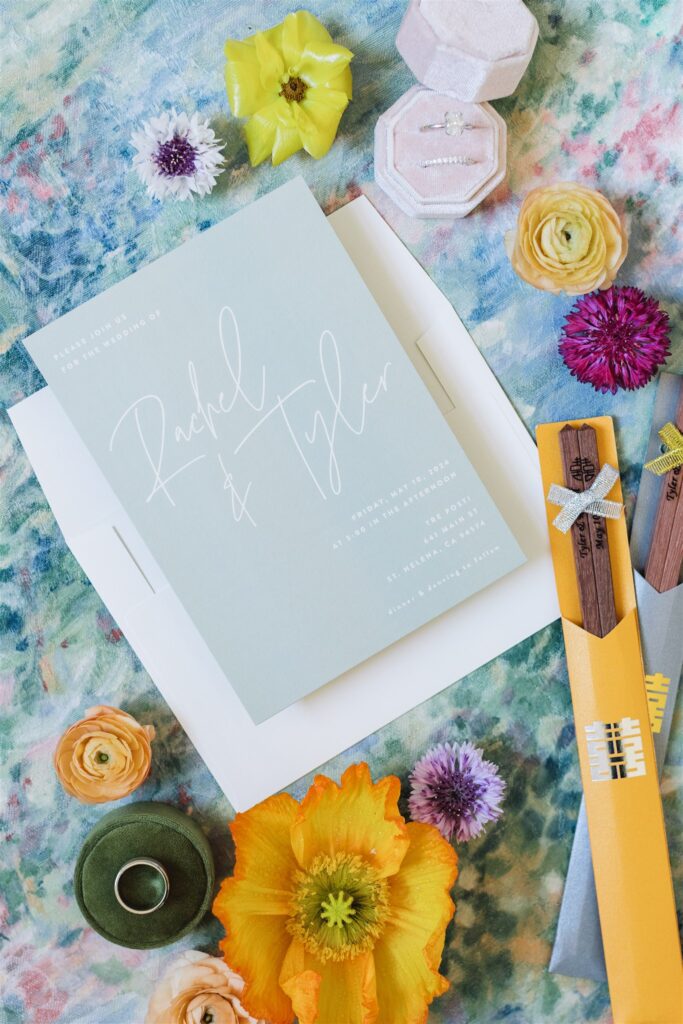 A Napa Valley wedding invitation for "Rachel & Tyler" is surrounded by colorful flowers, a green ribbon, rings in a box, and two bright yellow envelopes with wooden details on a floral background.