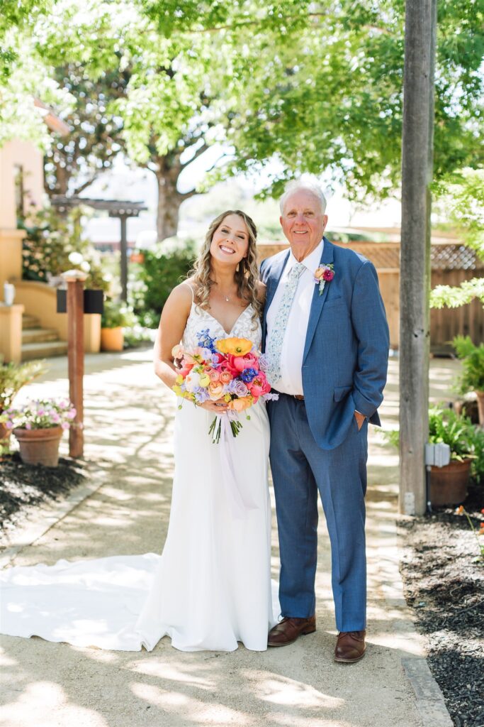 A bride and an older man in formal attire stand outdoors on a sunny Napa Valley day, smiling at the camera. She holds a colorful bouquet amidst the lush greenery.