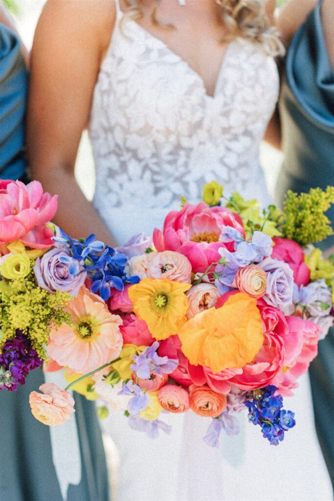 In a white lace dress, a person gracefully holds a vibrant bouquet filled with colorful blooms in pink, yellow, and purple—perfectly capturing the romance of a Napa Valley wedding.