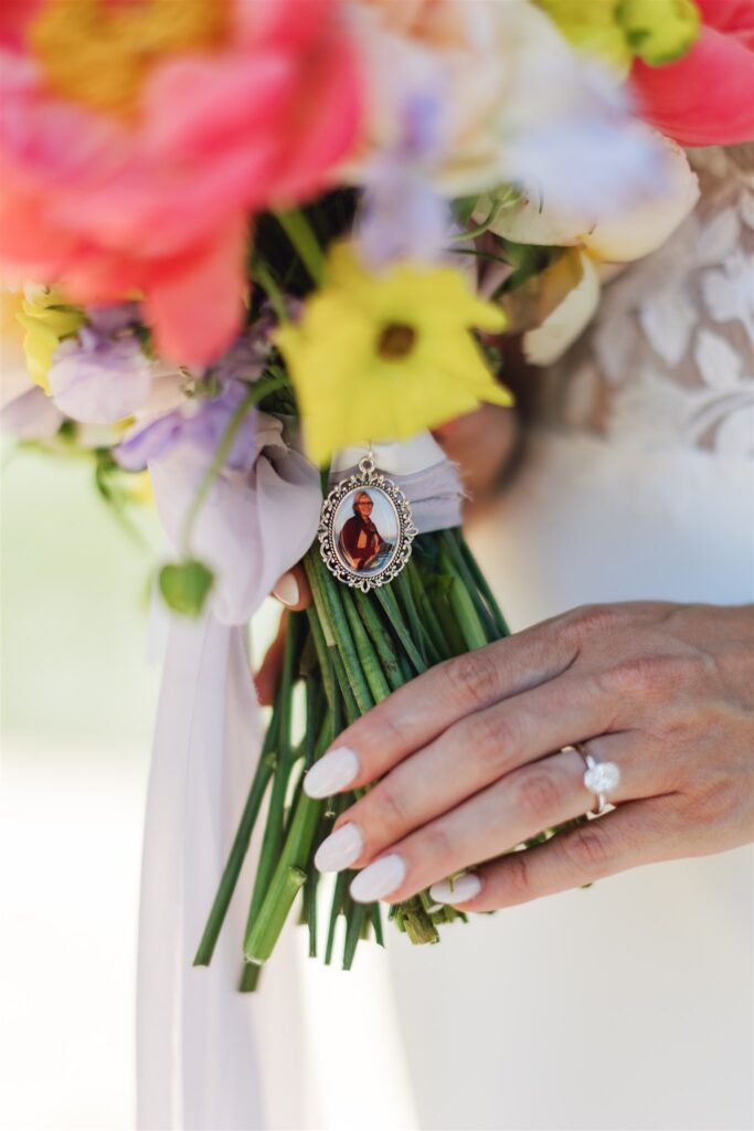 A hand with a ring holds a bouquet of colorful flowers, adorned with a cameo-style brooch featuring a person, capturing the timeless elegance of a Napa Valley wedding.