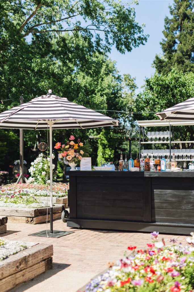Outdoor bar setup with striped umbrellas, various bottles, and glasses, perfect for a Napa Valley wedding. Surrounded by colorful flower beds and lush greenery, it offers an enchanting setting to celebrate love amidst nature's beauty.