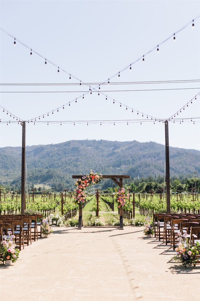 Outdoor Napa Valley wedding setup with wooden chairs and floral arrangements facing an arbor, set against a backdrop of vineyards and mountains under string lights.