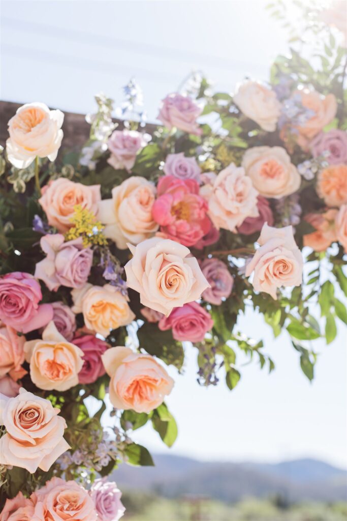 A colorful floral arrangement with pink, peach, and lavender roses graces the scene, reminiscent of a vibrant Napa Valley wedding beneath a bright, sunny sky.