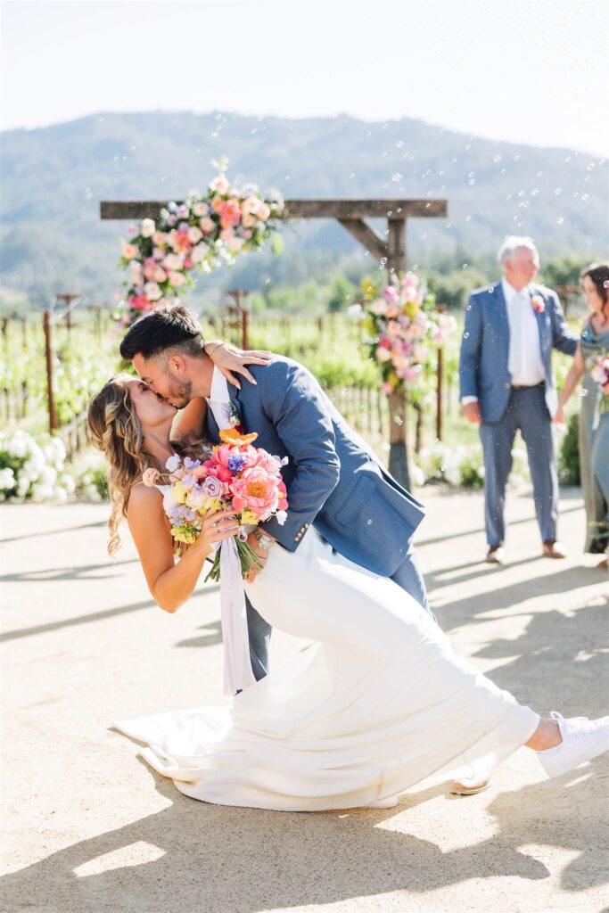 A couple in wedding attire shares a kiss beneath a floral arch, surrounded by onlookers and the breathtaking landscape of a Napa Valley wedding.
