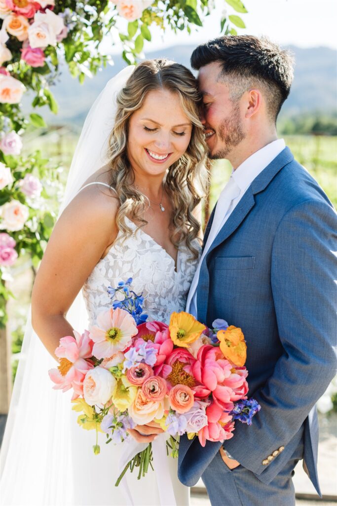 A bride and groom stand closely together outdoors, their love blooming as vibrantly as the colorful bouquet of pink, orange, and yellow flowers she holds. Surrounded by lush greenery and floral decorations, they capture the enchanting romance of a Napa Valley wedding.