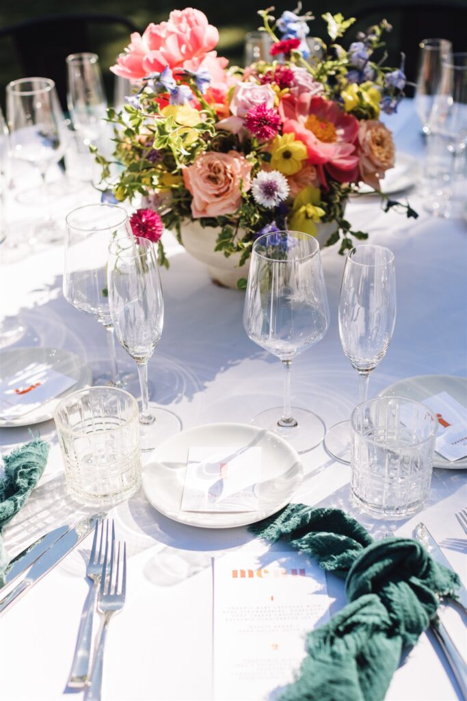 A white tablecloth with dishes, glassware, and green napkins sets the scene for a meal reminiscent of a Napa Valley wedding. A colorful floral arrangement is perfectly centered on the table.